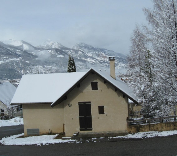 A quaint cabin is surrounded by snow-covered landscapes, with mountains visible in the background. The structure features a simple roof and a central door, complemented by a brick chimney and nearby trees. The pathway leads toward the entrance, enhancing accessibility.