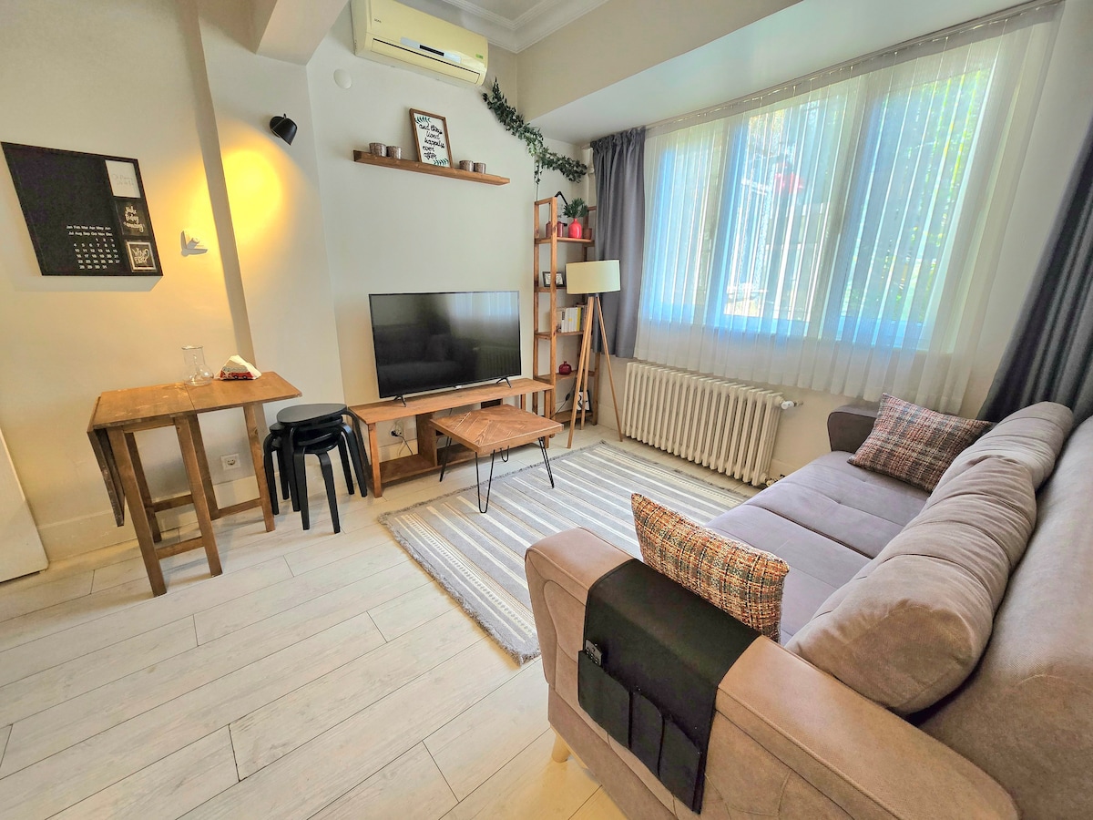 A cozy living area features a light-colored sofa with decorative cushions, providing a comfortable seating option. A flat-screen TV is positioned on a wooden stand, while natural light filters through sheer curtains. A small dining table is located nearby, accompanied by two black stools.