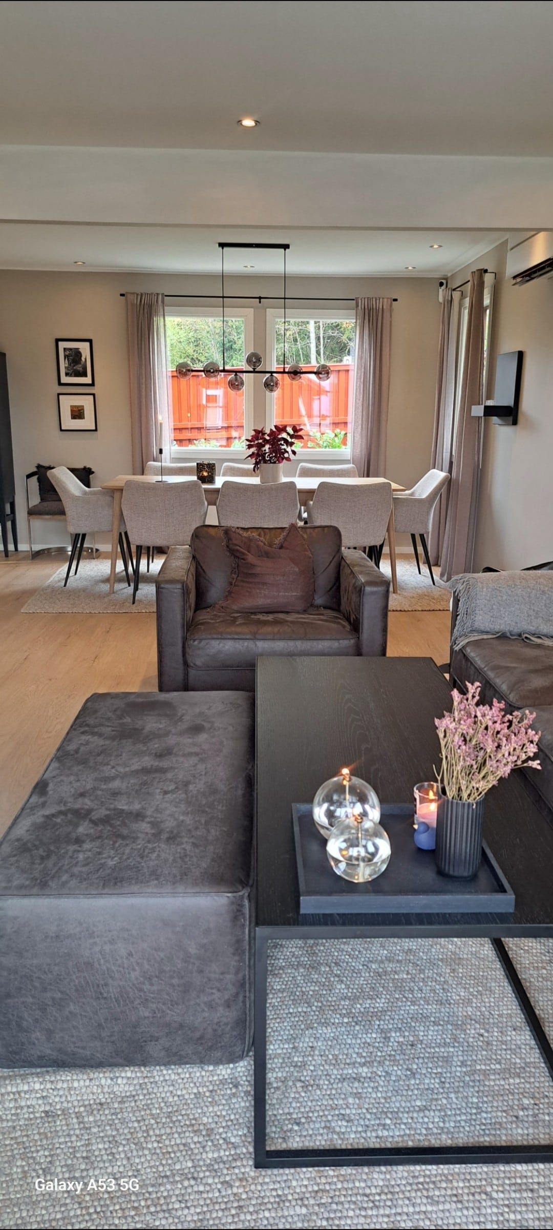 A modern living area features a comfortable seating arrangement and a sleek coffee table. Natural light enters through large windows, illuminating the space. Dining chairs are positioned around a rectangular table in the background, creating an inviting atmosphere for family gatherings.