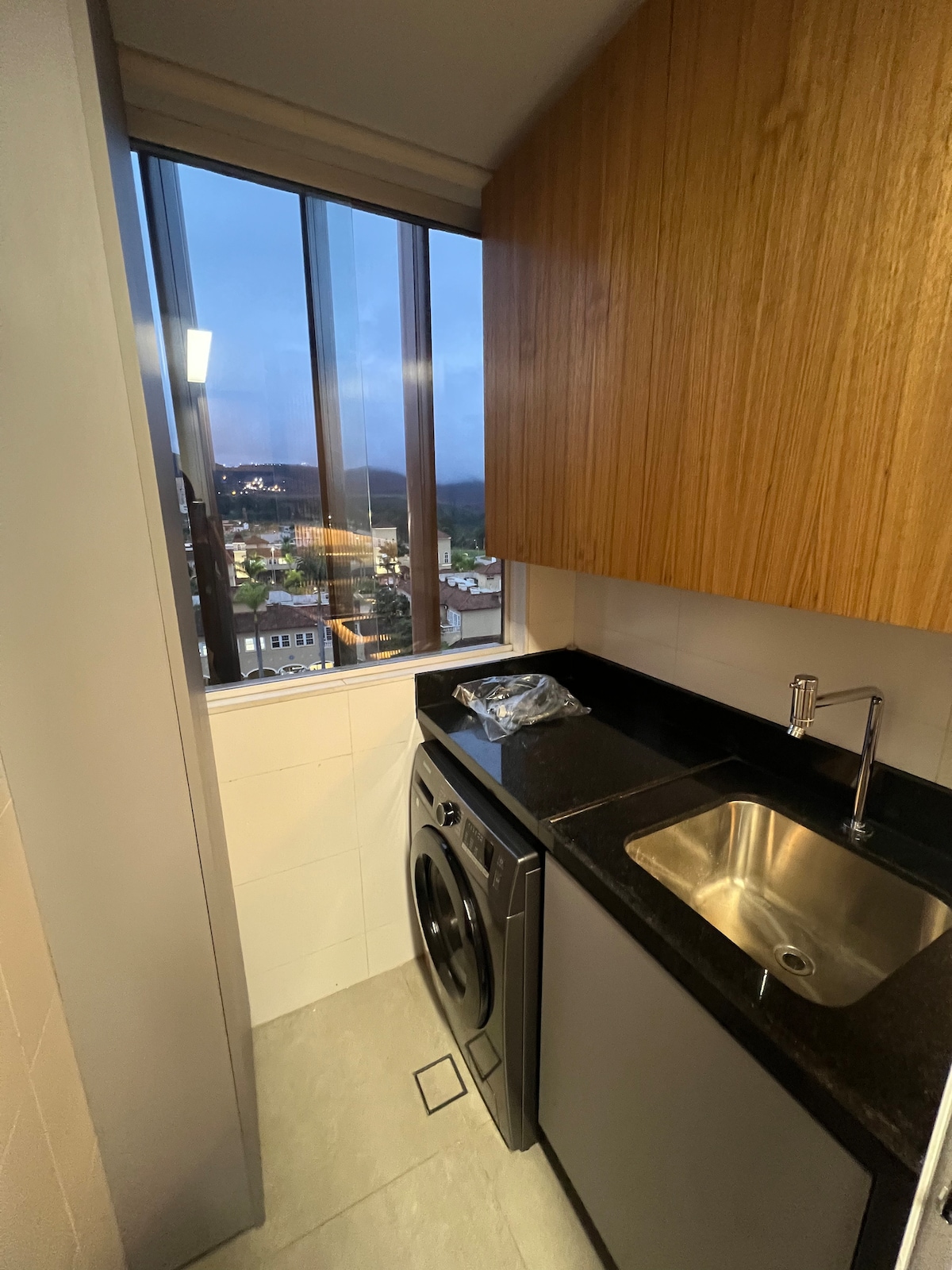A compact laundry area is visible, featuring a washing machine and a small stainless steel sink. Dark wooden cabinetry provides storage above while a large window allows natural light to enter, offering views of the surrounding landscape.
