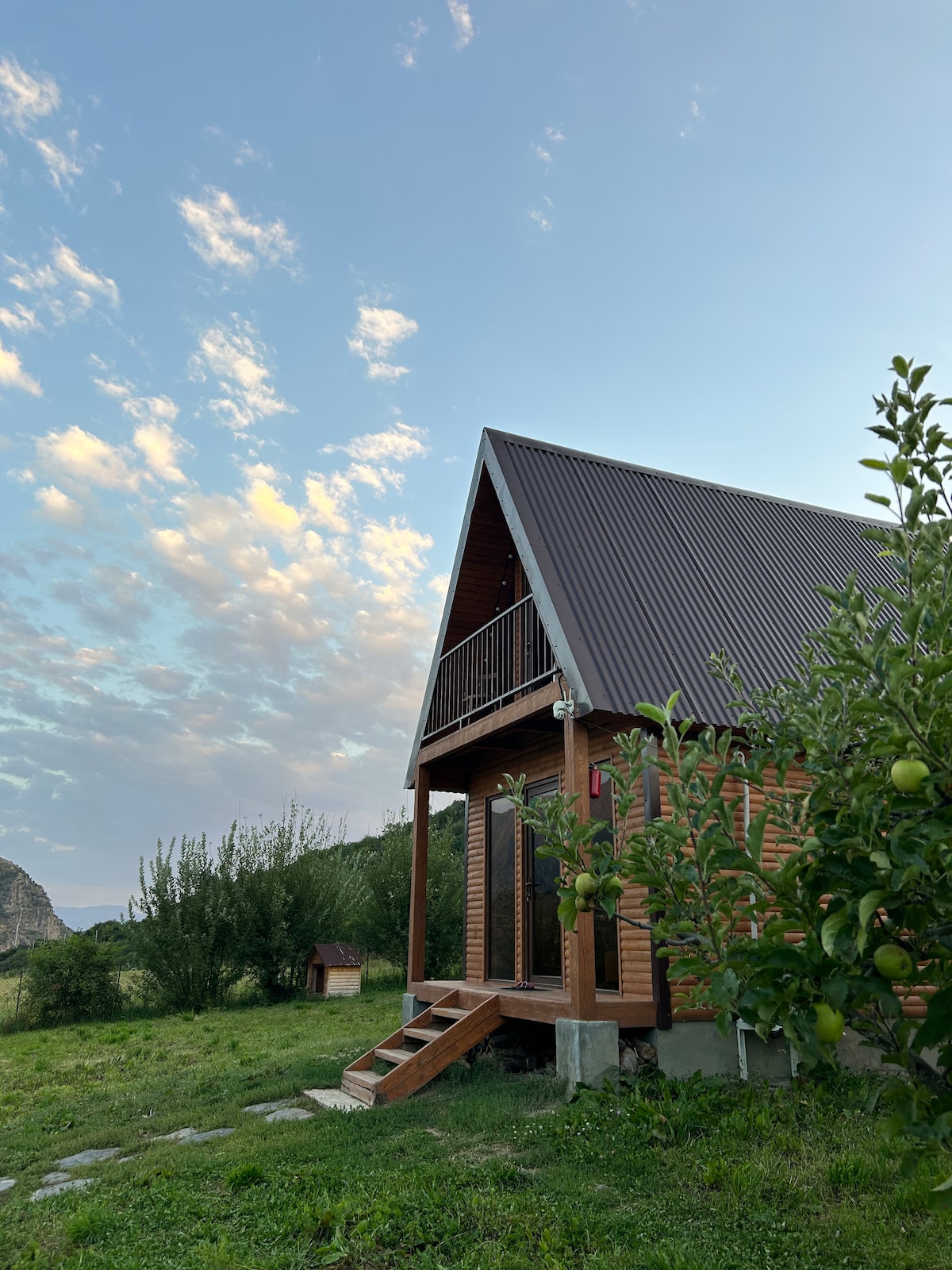 A modern wooden cabin is depicted with a sloped roof, surrounded by lush greenery. Steps lead up to a porch area, while trees with fruit are visible nearby. The sky is clear with scattered clouds, contributing to a serene outdoor setting.