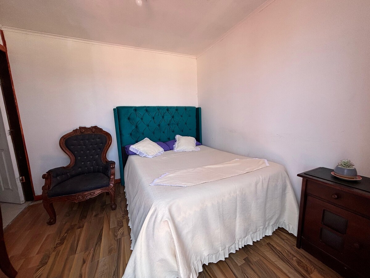 A spacious sleeping area features a tufted teal headboard above a neatly made bed. White linens are complemented by purple pillows. A dark wood nightstand sits beside the bed, and a vintage armchair is positioned nearby, enhancing the room's comfort.