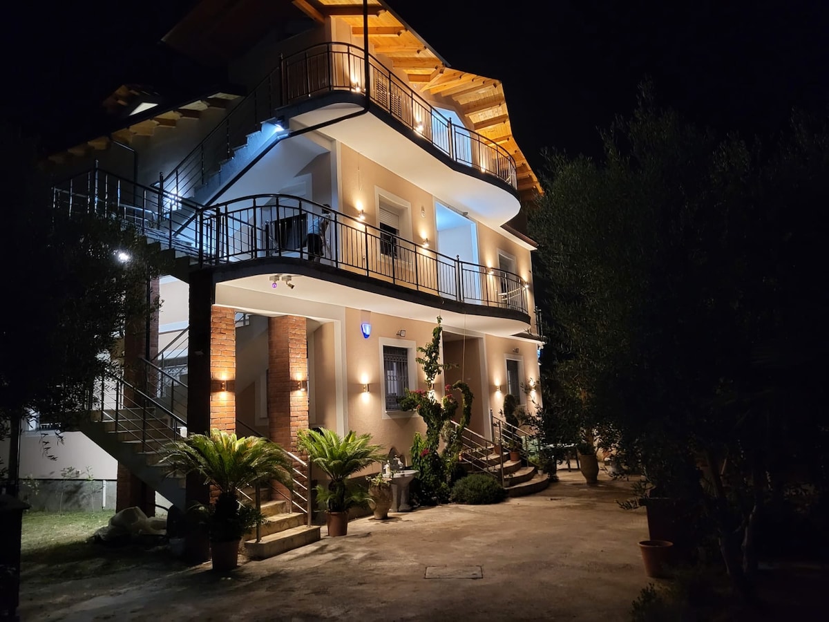 The modern three-story apartment building is illuminated at night, highlighting its architectural features. Balconies are visible on each level, surrounded by greenery and planters. The entrance is framed by steps leading to a spacious pathway, enhancing accessibility and inviting a pleasant approach.