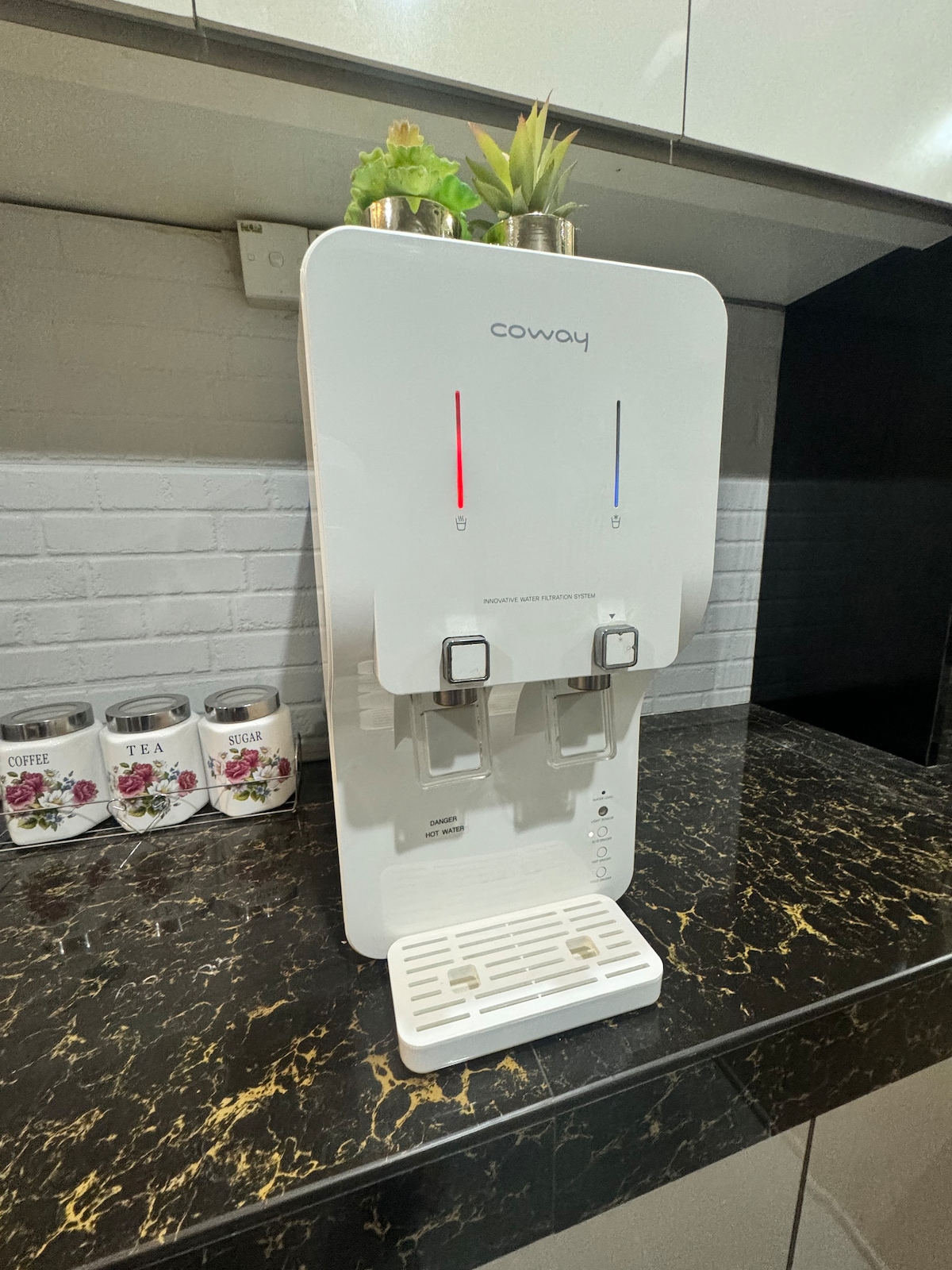 A water dispenser is positioned on a dark marble countertop, featuring two spouts for hot and cold water. Decorative plants are placed on top. Nearby, canisters labeled for coffee, tea, and sugar are neatly arranged.