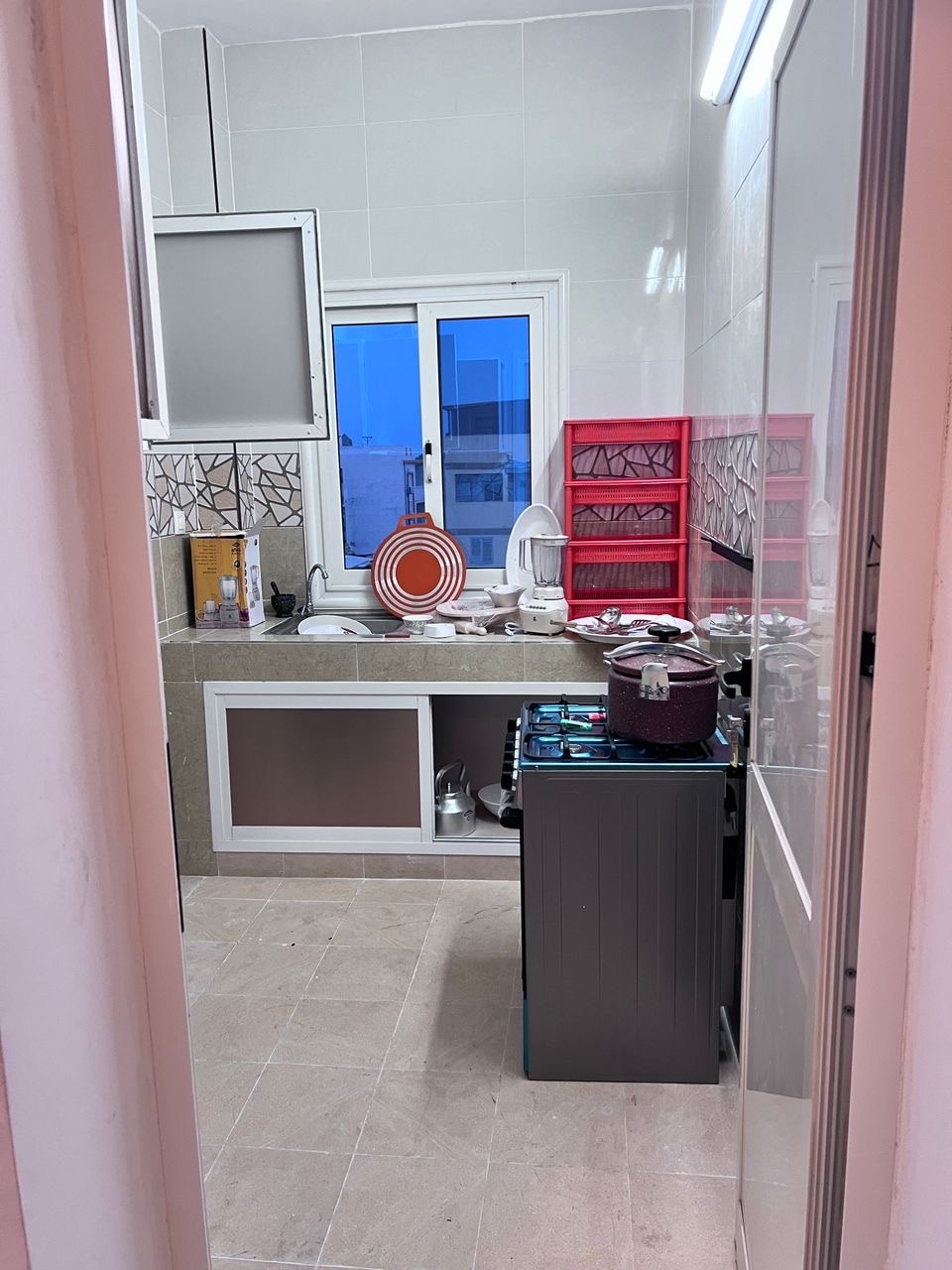 A functional kitchen is displayed, featuring streamlined cabinetry and a countertop. A window allows natural light to enter, with dishes organized on a red rack. A stove is positioned against the wall, and various utensils are visible throughout the space.