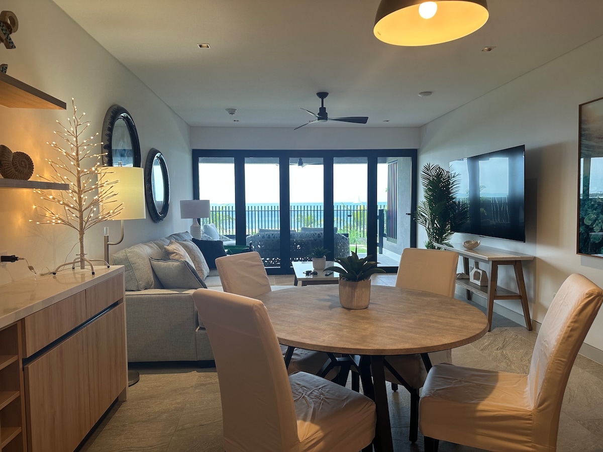 An open-concept living area showcases a circular dining table surrounded by four upholstered chairs. A light-colored sofa with accompanying cushions is positioned near large glass doors that provide panoramic views of the ocean. A television is mounted on the wall opposite, completing the modern aesthetic.