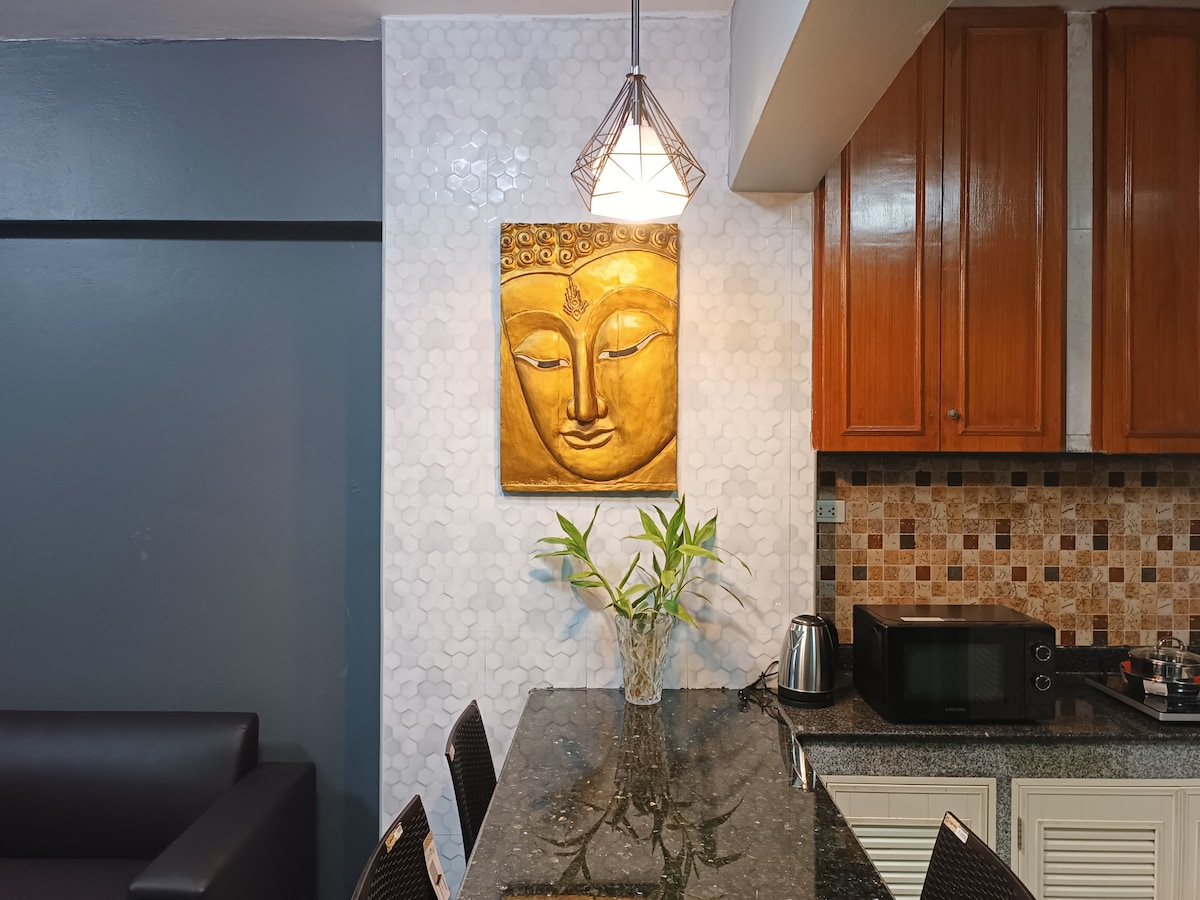 A well-lit kitchen area features a sleek granite countertop with a small vase of greenery. Above, a pendant light provides illumination, while wooden cabinets are mounted above the countertop. A microwave and an electric kettle are present, enhancing the functional appeal of the space.