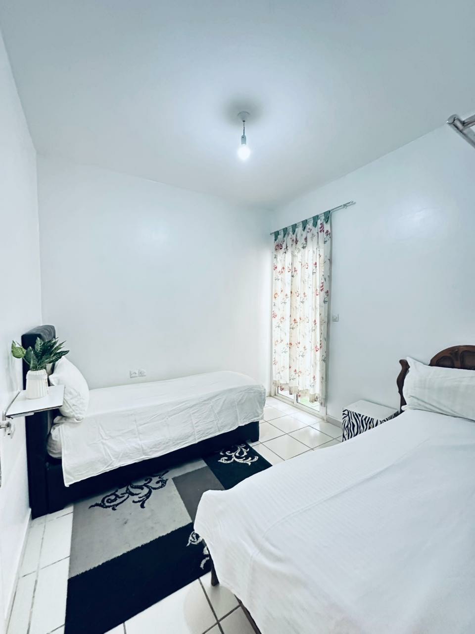 A bright and simple room features two spacious beds with white linens. A light curtain with floral patterns allows natural light to fill the space. A small table with a plant sits beside one bed, and decorative rugs are visible on the tiled floor.