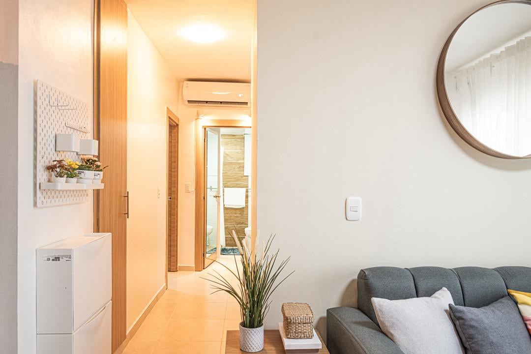 A light-filled entryway is featured, showcasing a gray sofa adorned with several decorative cushions. A round mirror reflects the space, while greenery adds a touch of nature. The doorway at the end leads to a bathroom area, creating a welcoming environment.