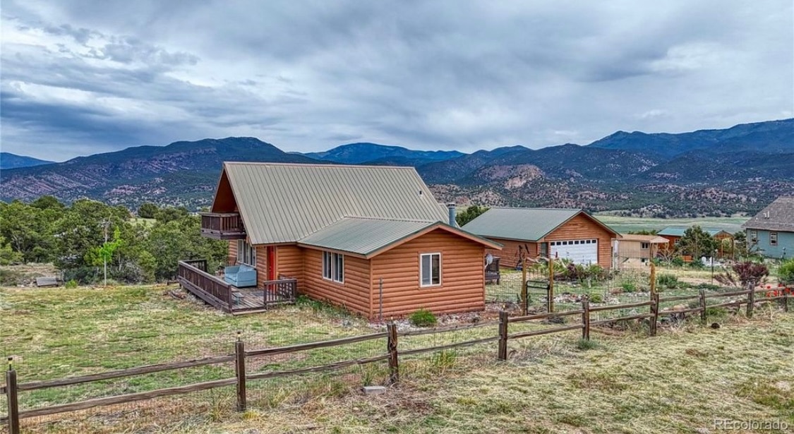 A spacious lodge-style home is set against the backdrop of rolling hills and the dramatic mountain range. The exterior features a combination of wooden siding and a metal roof. Surrounding greenery includes patches of grass and a wooden fence delineating the property.
