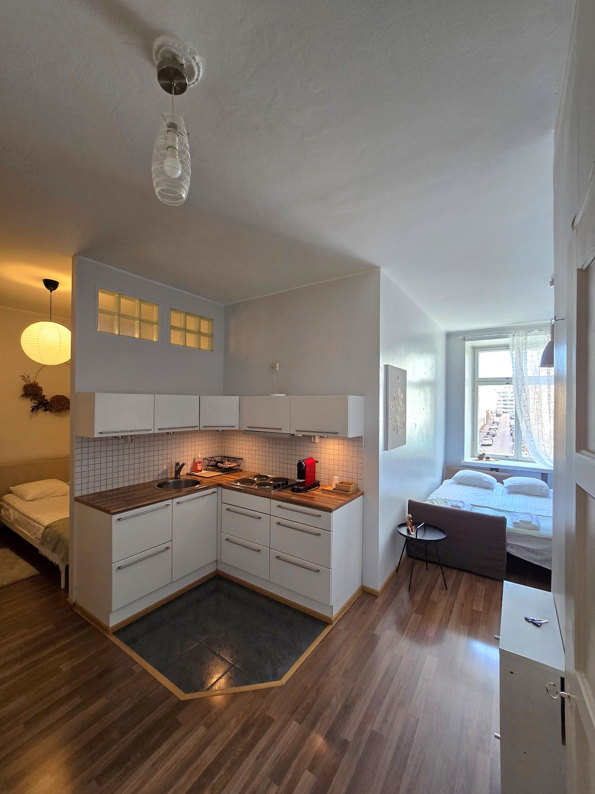 An open-plan studio features a compact kitchen with modern appliances, highlighted by an L-shaped countertop. Natural light enters through a nearby window, illuminating a cozy sleeping area with two beds positioned adjacent to each other. Neutral tones create a calm environment throughout the space.