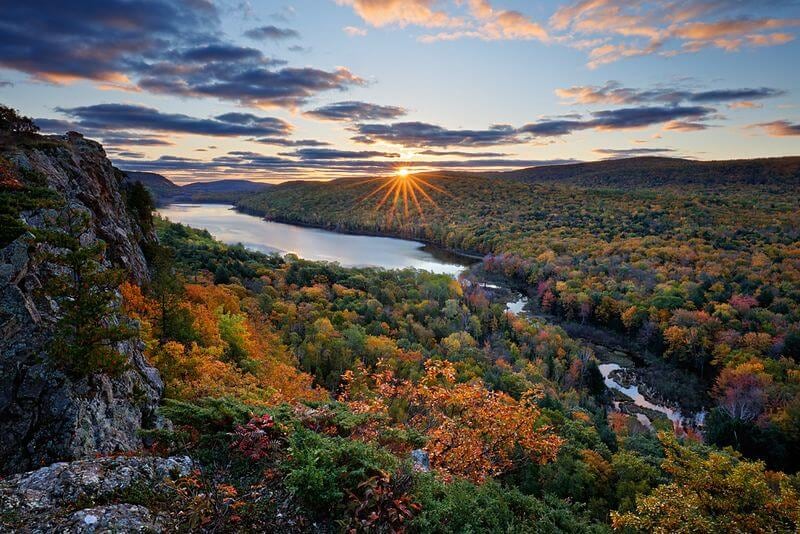 The image captures a panoramic view of a river winding through a vibrant autumn forest. Sunlight breaks through clouds, creating a radiant starburst effect over the horizon. The landscape is adorned with a rich palette of orange, yellow, and green foliage, set against a serene backdrop.