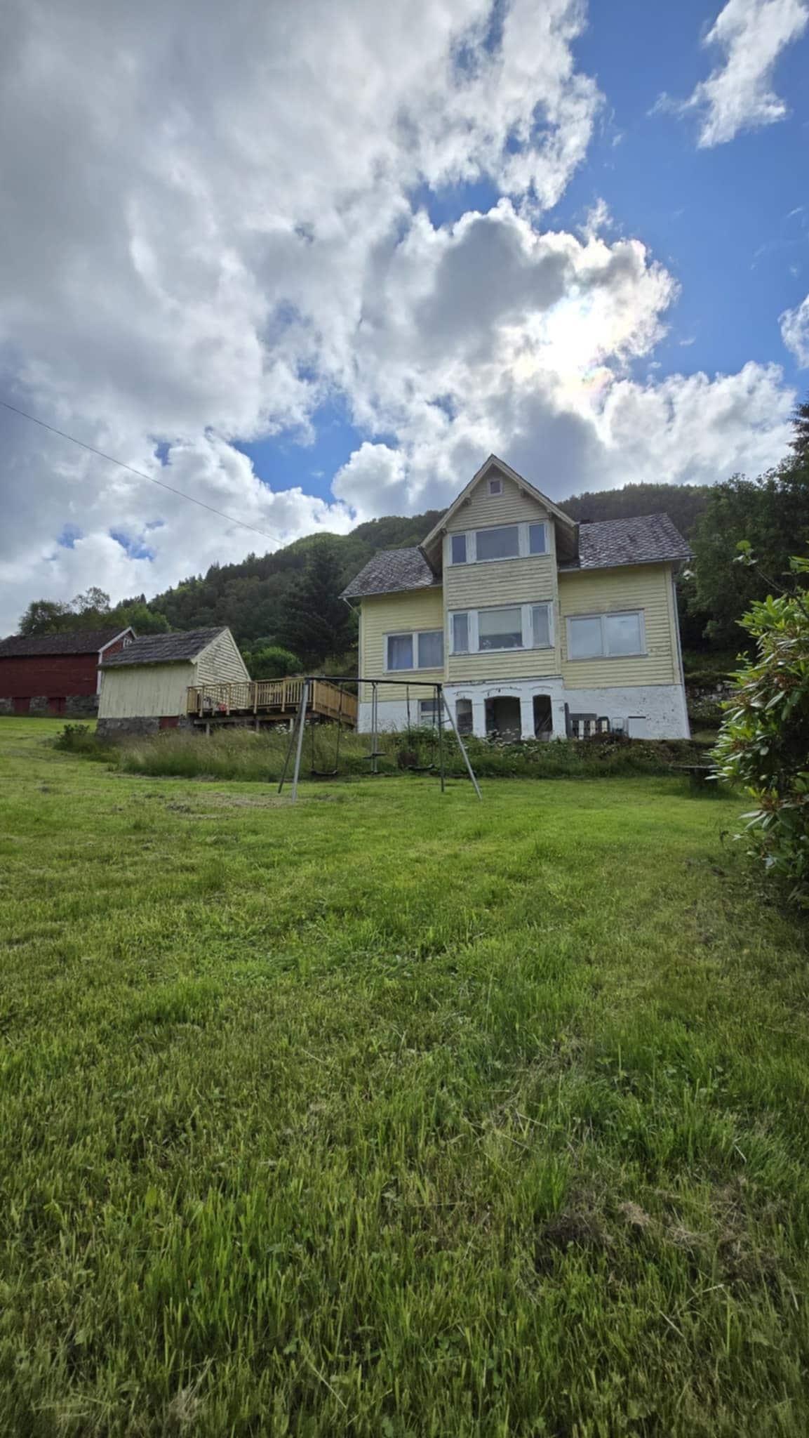 A charming two-story house is situated in a grassy area, surrounded by trees and hills. The facade, painted in soft yellow, features multiple large windows that allow natural light to enter. Nearby structures, including a red building, are visible in the background beneath a partly cloudy sky.