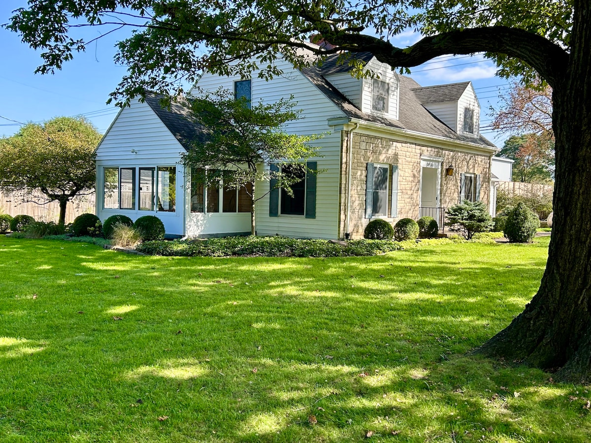 The exterior of a Cape Cod home is showcased amidst a lush green lawn. Sunlight filters through trees, illuminating the inviting sunroom that features large windows. The property is bordered by manicured shrubs, adding a touch of greenery to the charming facade.