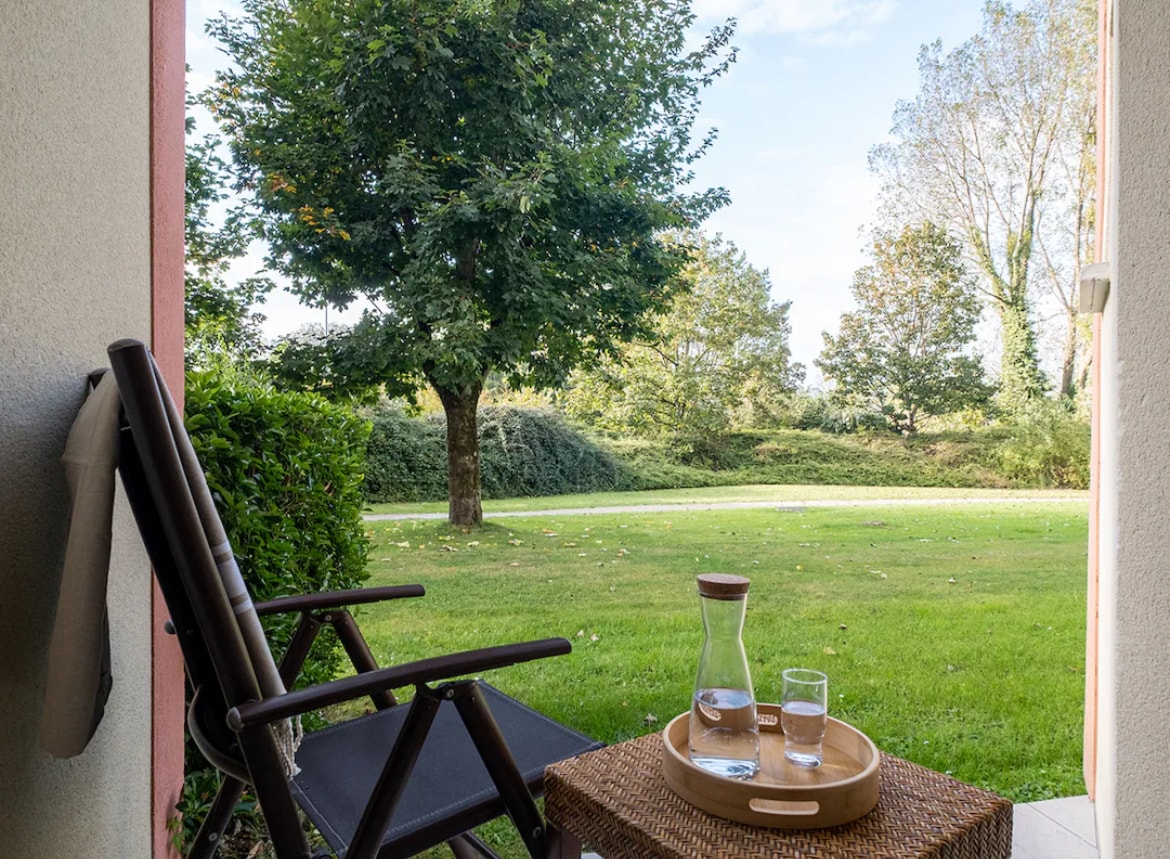A comfortable outdoor seating area features a wooden chair positioned on a small patio. A tray with a glass of water and a carafe is placed nearby. Lush green grass and a tree are visible in the serene garden view beyond.