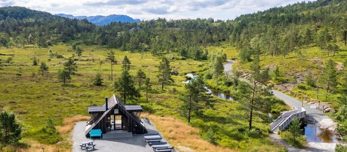 An aerial view captures a modern cabin surrounded by lush greenery and open meadows. A winding path leads through the landscape, while outdoor seating areas are visible in front of the cabin. Hills and trees frame the horizon, enhancing the serene setting.