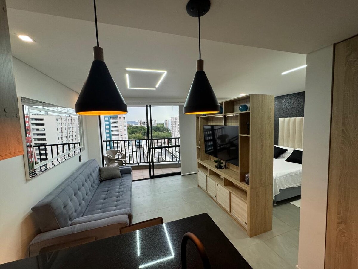 A modern living area features a comfortable gray sofa and a wooden entertainment unit containing a television. Large glass doors open to a balcony, allowing natural light to illuminate the space while city views are visible. Sleek pendant lights hang above an adjacent dining area.