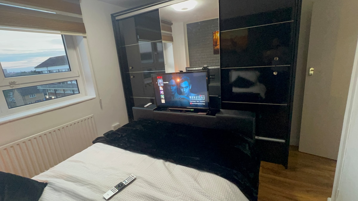 A compact bedroom space is showcased, featuring a spacious bed layered with a dark blanket. A flat-screen television is mounted on a contemporary entertainment unit, while light filters through the window, illuminating the dark cabinetry and wooden floor.