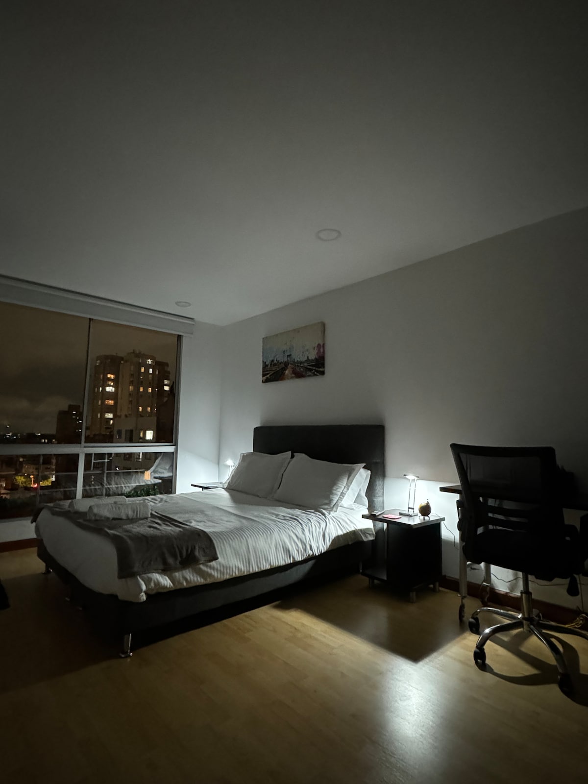 A spacious bedroom is depicted with a large bed, featuring crisp white linens and a dark headboard. A desk with an office chair is positioned nearby, and a soft light emanates from a bedside table lamp. City lights can be seen through the window, adding ambiance.