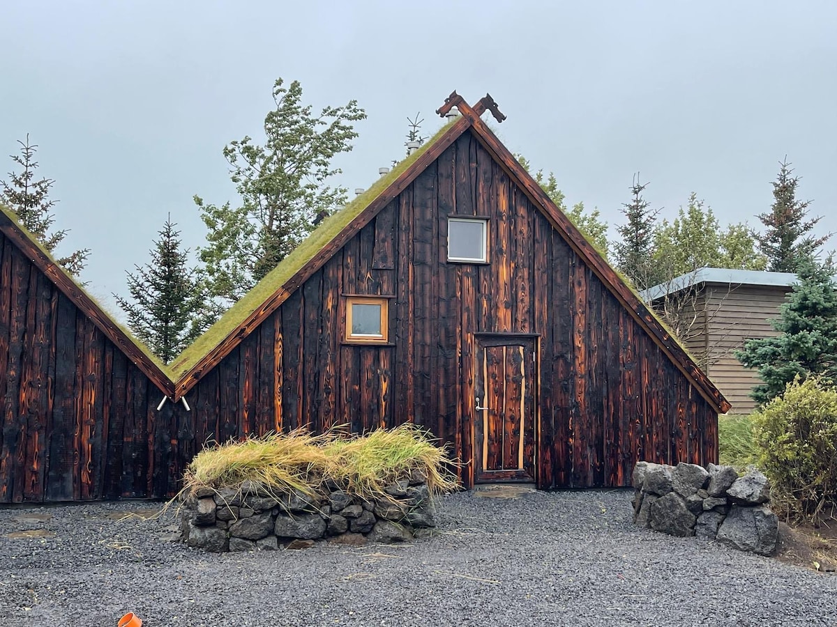 A rustic wooden cabin exhibits a characteristic A-frame design, featuring a dark wood exterior. Small windows are visible, allowing for natural light to enter. A garden area with low greenery is present in front, along with rocky landscaping that adds to the cabin's charm.