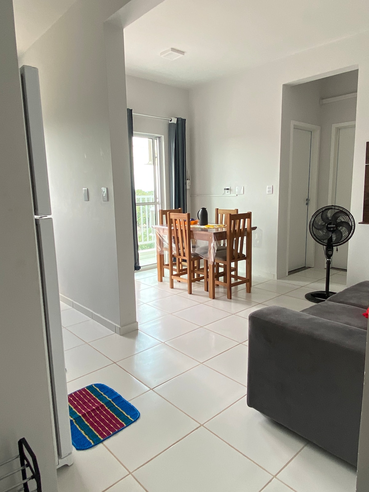 A dining area features a wooden table surrounded by four chairs, with light entering through a large window. A small fan is positioned near the corner, while a gray sofa is visible nearby. The flooring is tiled, and walls are painted in light tones.