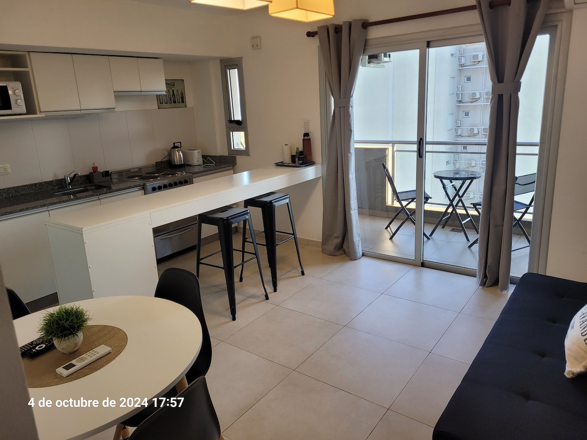 A contemporary kitchen features a spacious countertop and modern appliances. Bar stools are positioned at the counter, while a dining table is set with minimal decor. Large sliding doors allow natural light and provide access to a balcony with a small outdoor seating area.