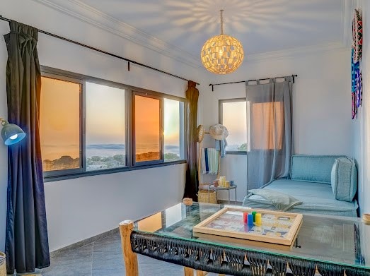 A cozy seating area is highlighted by large windows revealing a serene sunset over the ocean. Light filters through sheer curtains, and a decorative woven pendant light adds a soft glow. A game board rests on a glass-topped table, surrounded by comfortable seating.