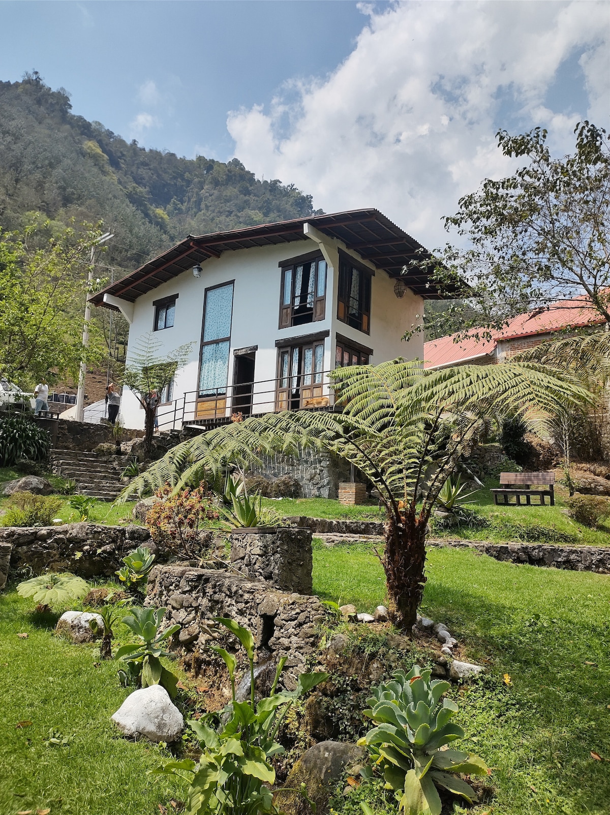 A two-story house is situated against a backdrop of lush greenery, showcasing a blend of architectural styles. Surrounding garden spaces feature a variety of plants and ferns, while stone pathways lead to the entrance. Mountains rise majestically in the distance under a partly cloudy sky.