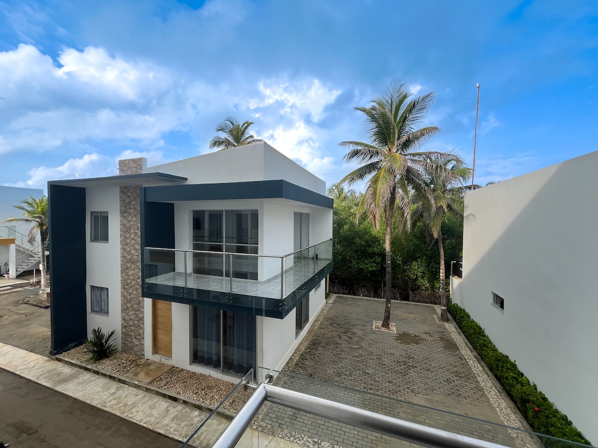 The two-story building features a modern design with large glass balconies and a stone accent wall. Surrounding palm trees create a relaxing environment. A well-maintained parking area is visible, complemented by a neatly paved path and greenery lining the property.