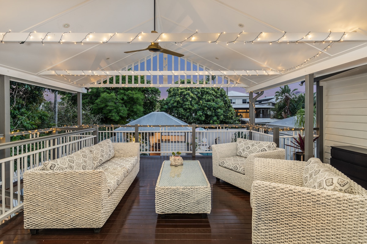 An outdoor lounge area features four comfortable seating arrangements made of woven material. A glass-top coffee table sits at the center, surrounded by warm, ambient lighting strung above. Lush greenery and rooftops are visible in the background, adding to the inviting atmosphere.