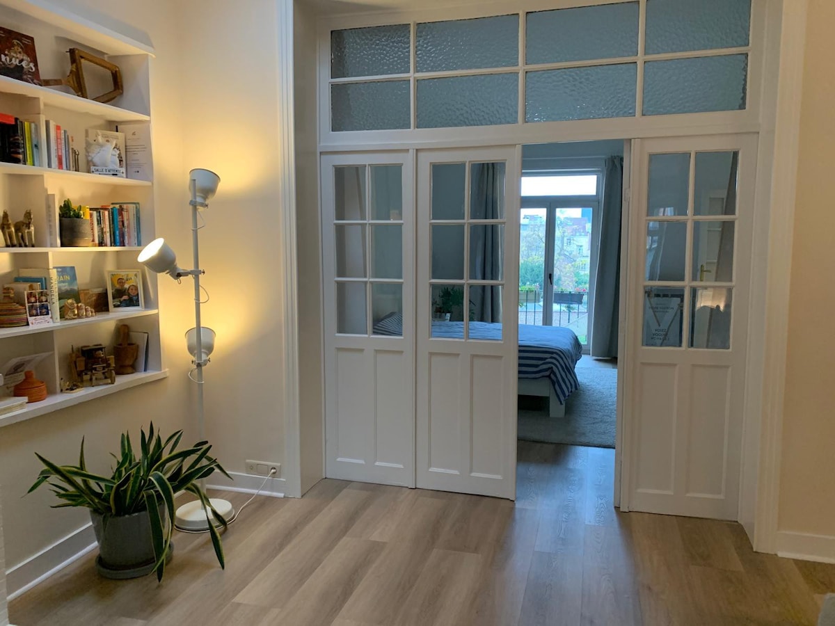 An entryway is depicted, featuring white double doors that separate the living area from a bedroom. A bookshelf with various decorative items lines one wall, while a modern floor lamp stands nearby. Light from the open balcony illuminates the space, highlighting smooth wooden flooring.