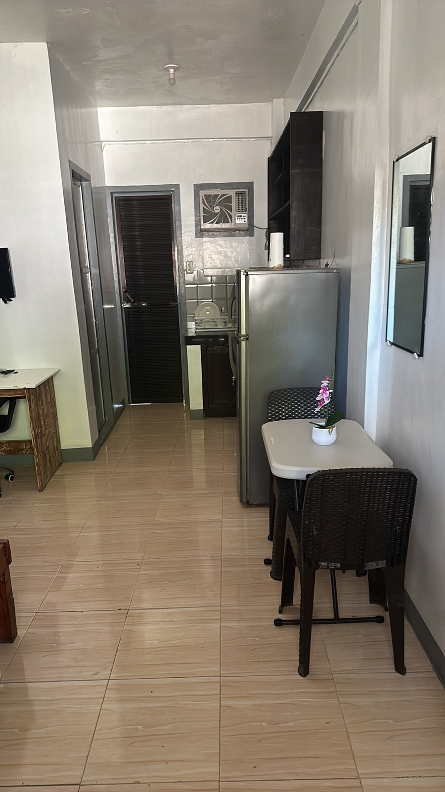 A compact kitchen area is visible, featuring a stainless steel refrigerator and a small dining table with two wicker chairs. A plant sits on the table, adding a touch of greenery. Light tile flooring extends throughout the space, creating an open and airy feel.