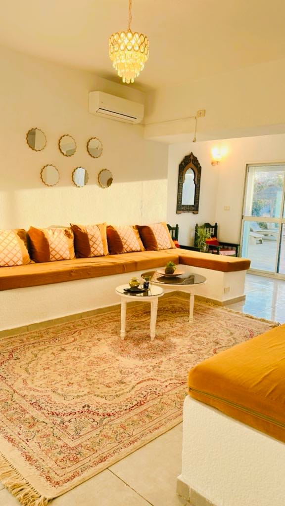 A bright living area features a comfortable seating arrangement with plush cushions against a white wall, complemented by decorative mirrors. A small coffee table is placed on a patterned rug, while natural light enters through a nearby sliding door, illuminating the space.