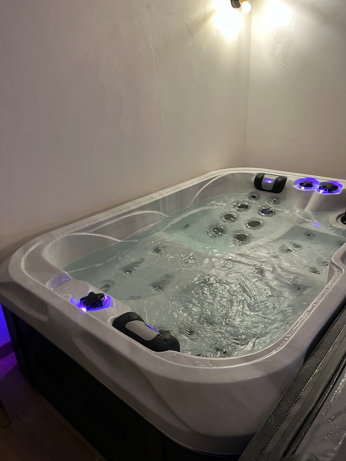 A private jacuzzi is displayed, featuring multiple jets and glowing blue LED lights. The interior of the tub contains bubbling water, creating a relaxing ambiance. The surrounding area is minimal, with walls partially illuminated to enhance the spa-like setting.