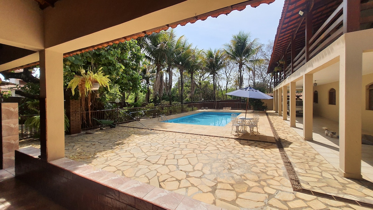 The outdoor area features a spacious pool surrounded by a stone deck. Tropical plants and palm trees provide a natural backdrop. A sunshade covers a seating arrangement, enhancing the space for relaxation. The layout allows for easy access to both the pool and adjacent covered areas.