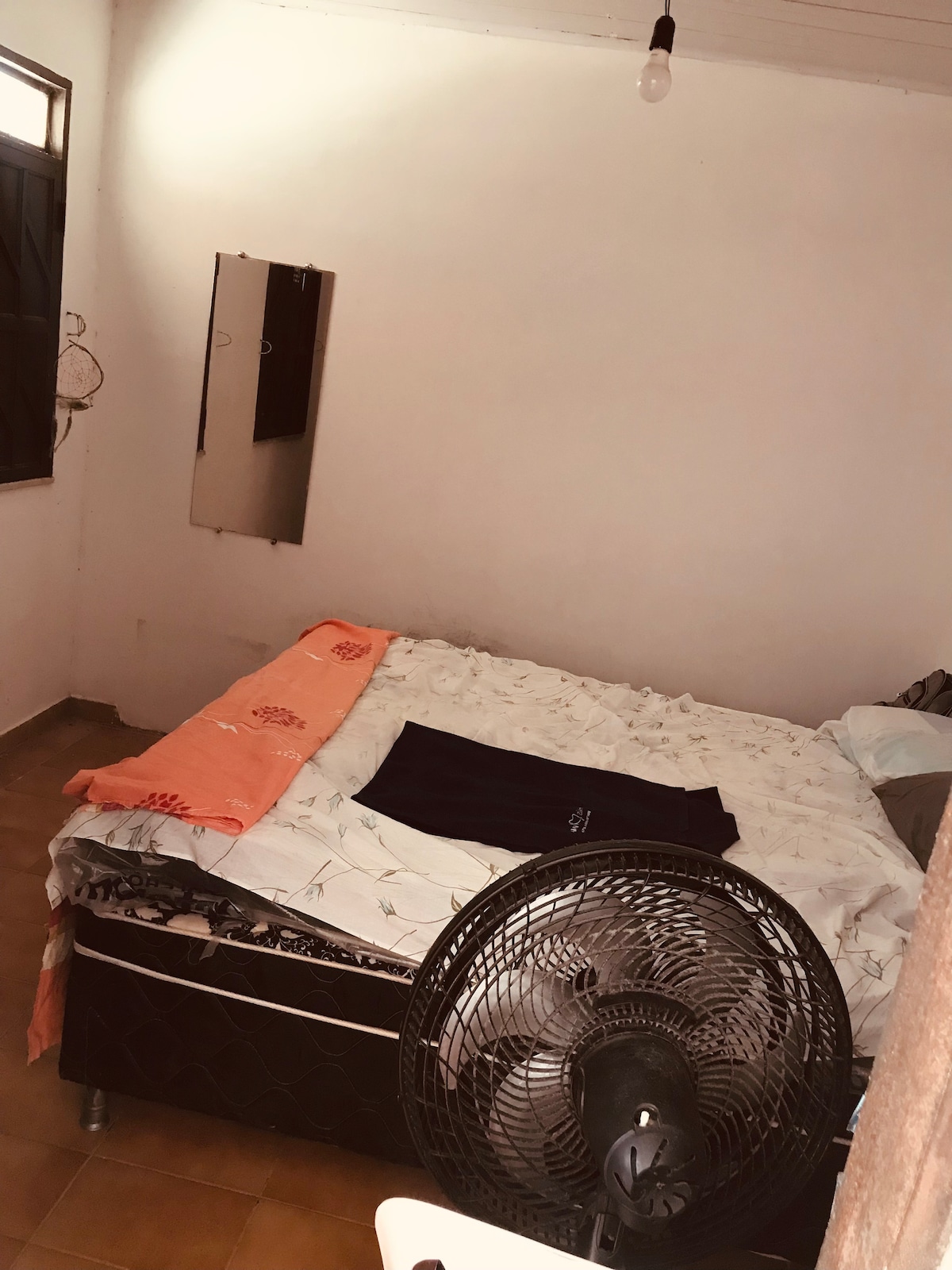 A simple bedroom features a spacious bed with light bedding and an orange throw blanket. A fan is positioned in the foreground, and a mirror is mounted on the wall. Natural light enters through a window, enhancing the uncluttered space.
