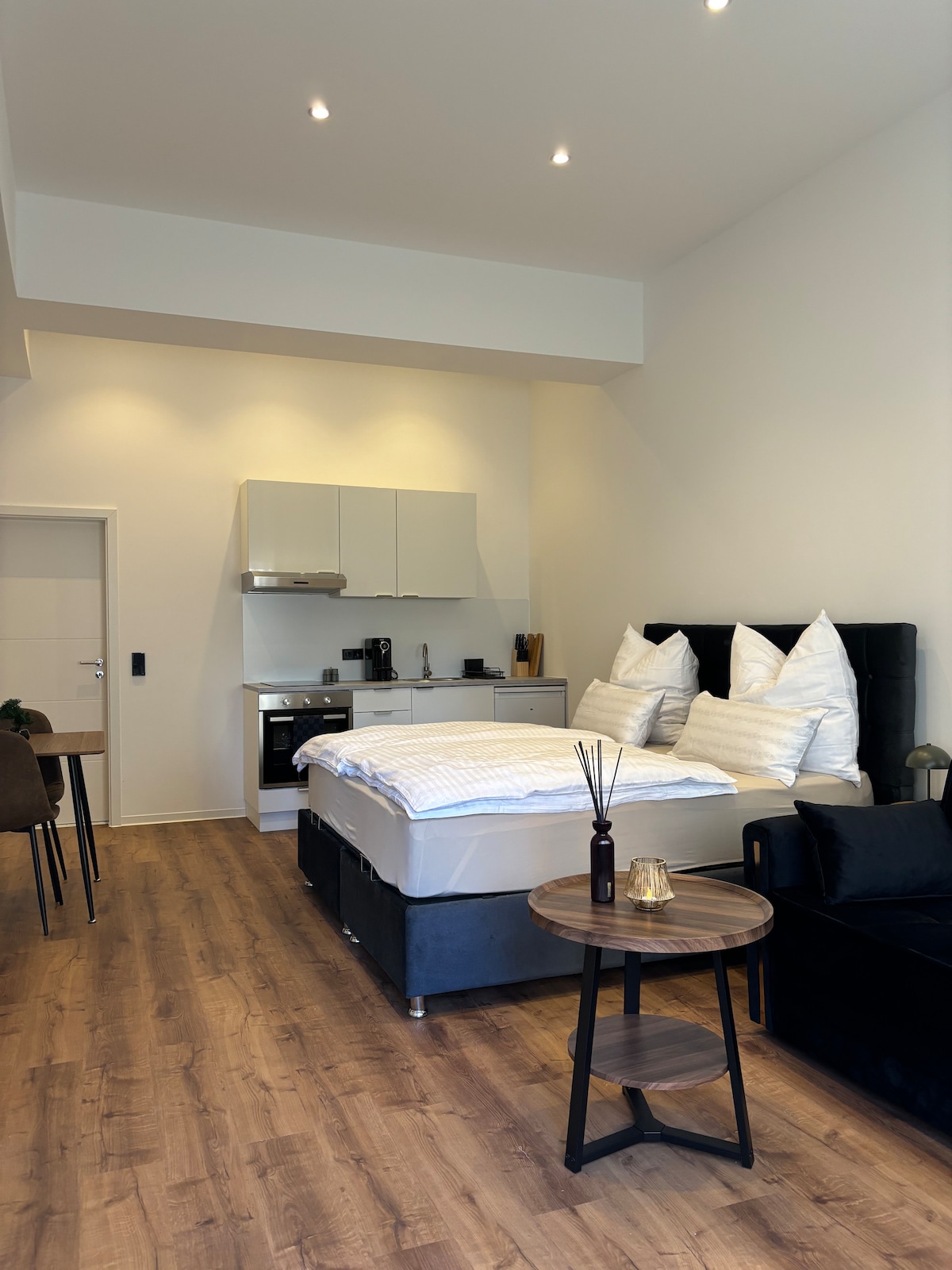 A spacious studio apartment features a comfortable bed with multiple decorative pillows. A kitchenette is visible, equipped with modern appliances. A small dining table and chairs are positioned nearby, while a cozy seating area is seen with a dark-colored couch. Natural light illuminates the wood floor.