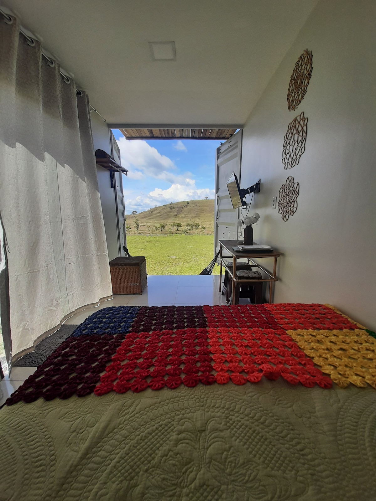 A cozy sleeping area is presented with a colorful, textured bedspread featuring various shades. A small desk and chair are positioned nearby, while large curtains frame the open view towards the landscape, allowing natural light to enter the space.