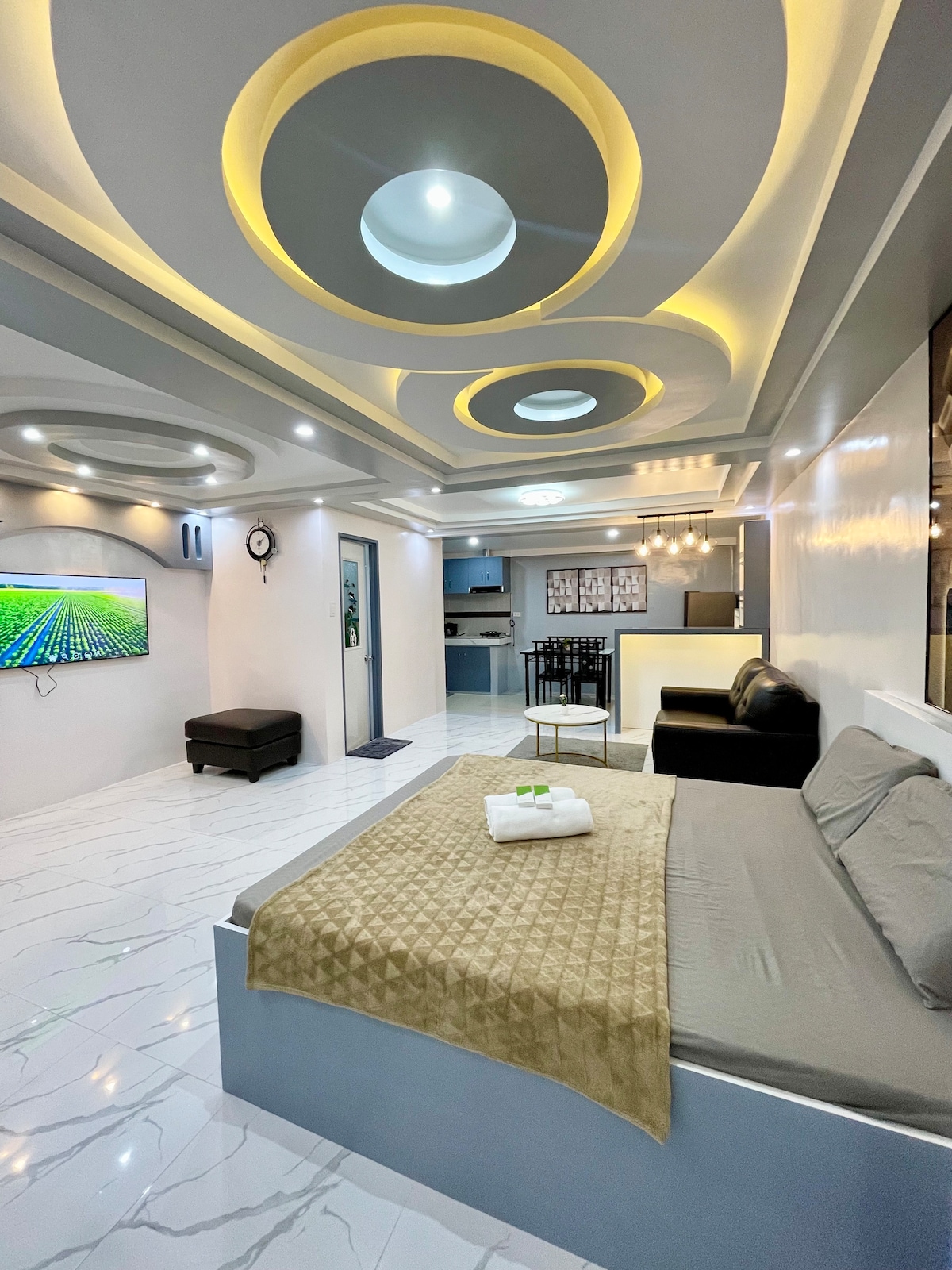The living area features a spacious bed with a beige throw blanket, complemented by soft couches. A flat-screen television is mounted on the wall, and a dining area with a four-seat table is visible. The ceiling is highlighted with circular designs and integrated lighting.