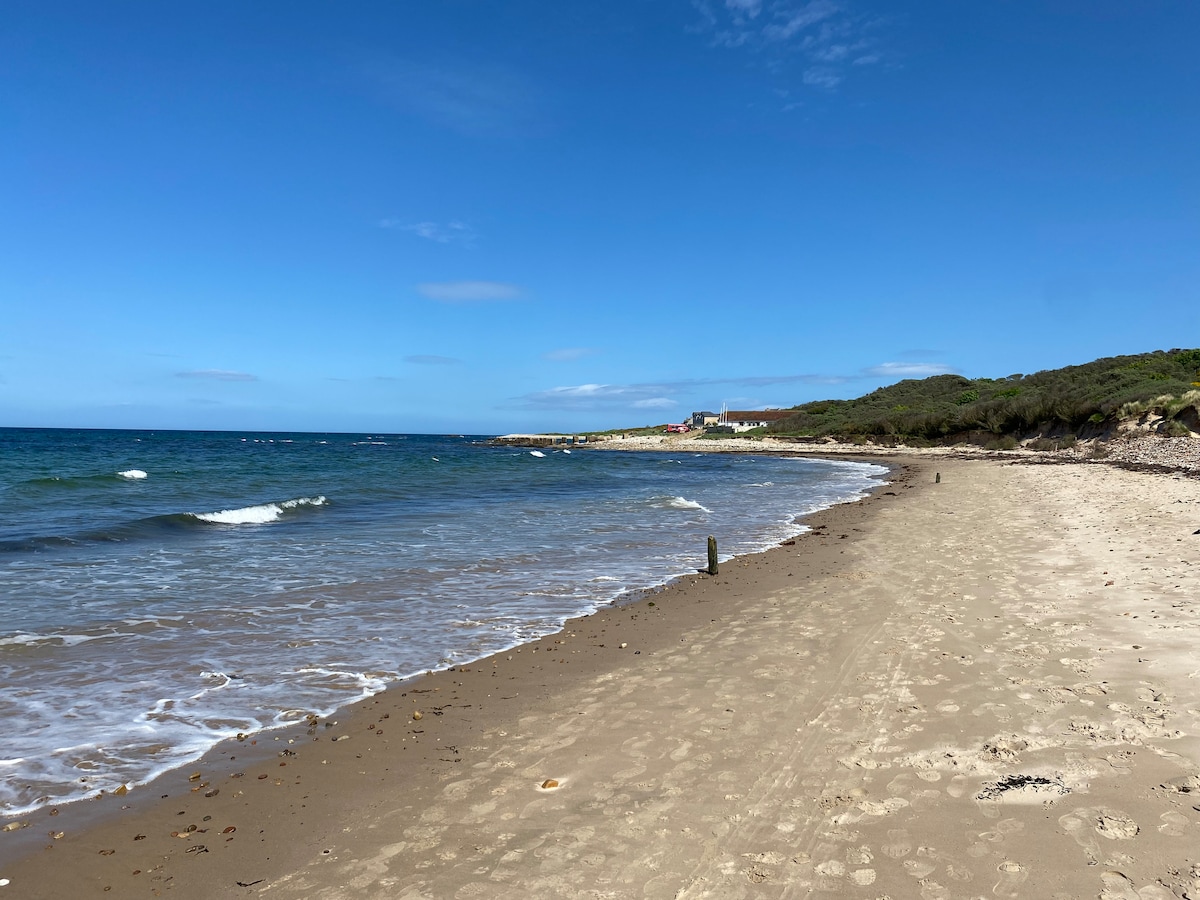 A sandy beach extends along the shoreline, meeting calm waves under a clear blue sky. Sparse vegetation is visible at the edge of the beach, with distant buildings framed against the horizon, contributing to a serene coastal landscape.