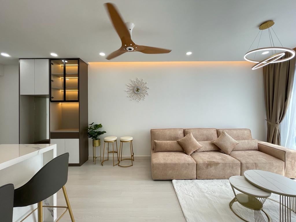 The living area features a comfortable sofa with decorative cushions, accompanied by elegant side tables. A modern ceiling fan is present, and the space is illuminated by soft lighting. The decor includes a wall accent piece, creating a stylish yet relaxed atmosphere.