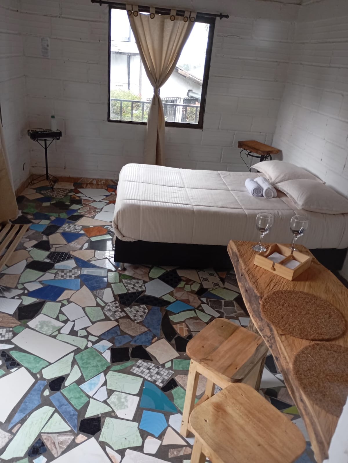 A cozy bedroom contains a spacious bed dressed with fresh linens. Beside the bed, small wooden tables provide functionality. A large window allows natural light to fill the room, accentuating the unique tiled flooring made of varied colors and textures.