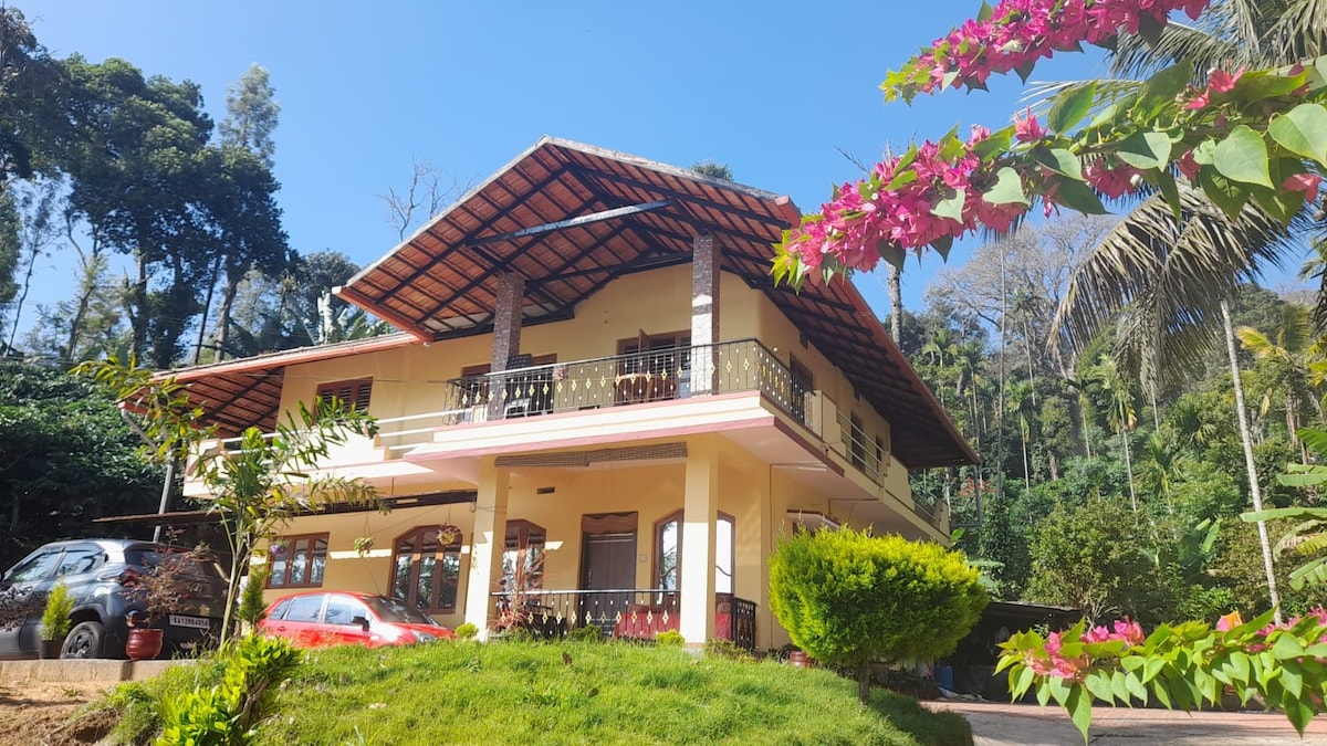 A two-story house is surrounded by lush greenery, reflecting a calm atmosphere. The structure features a shaded balcony with wrought-iron railings, while flowering plants add vibrant color to the exterior. A pathway leads to the entrance, enhancing accessibility to the inviting outdoor space.