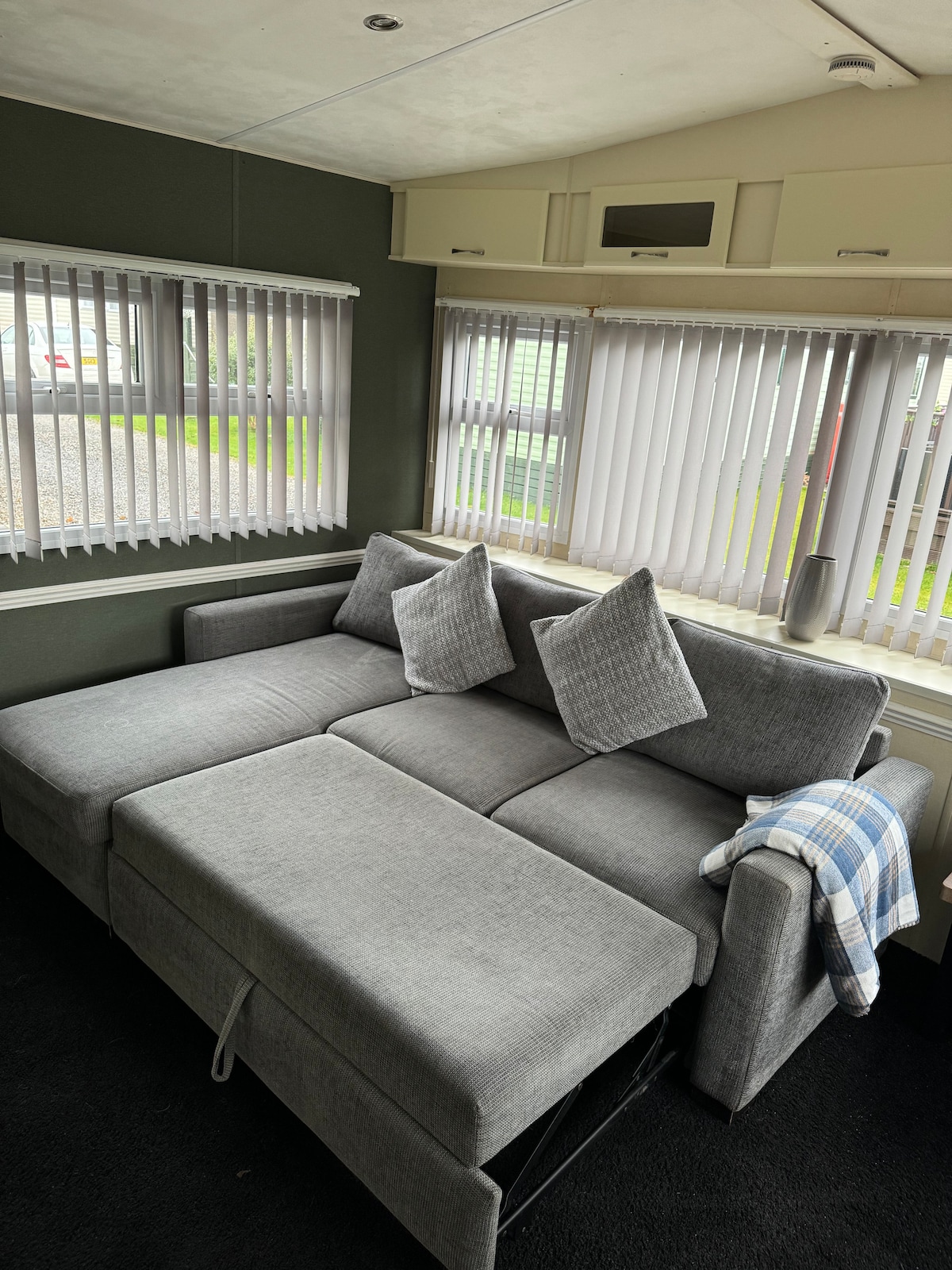 A modern gray sofa is positioned in a well-lit corner of the living area, featuring a pull-out section and two accent pillows. Light filters through the vertical blinds covering the windows, while a soft throw is casually draped over the armrest.