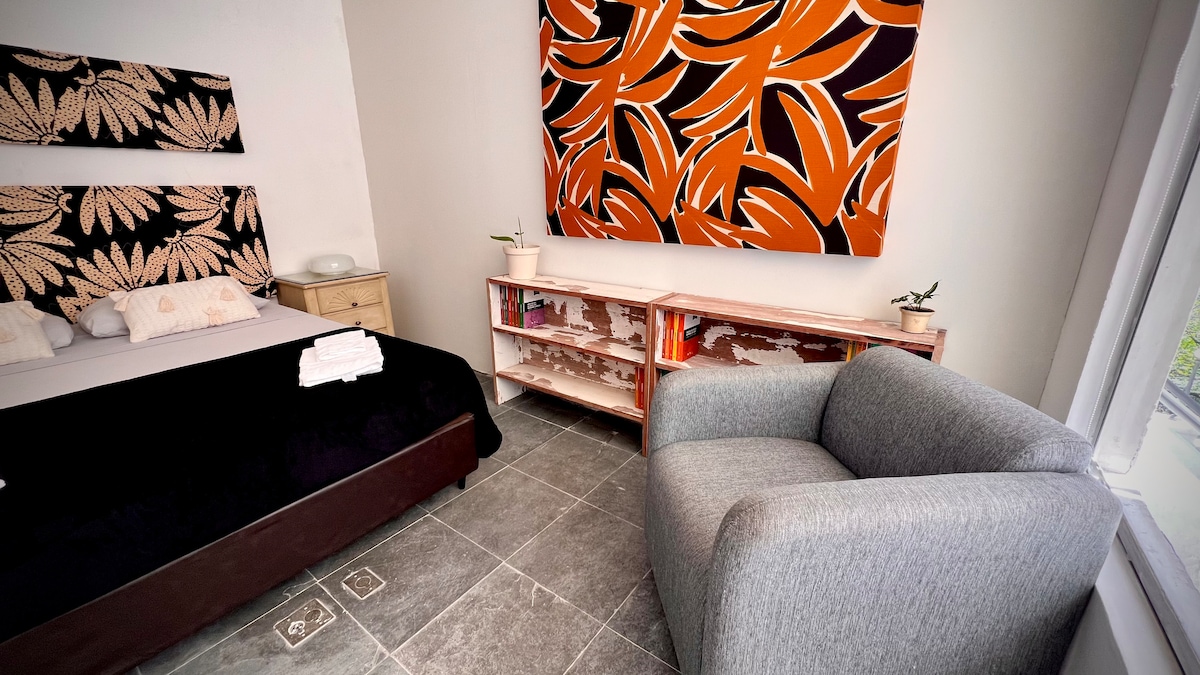 A cozy sleeping area is featured, with a spacious bed adorned with neutral-toned linens. A modern armchair is positioned nearby, and a stylish shelf with books pulls focus against the wall. Large windows allow natural light to illuminate the room.