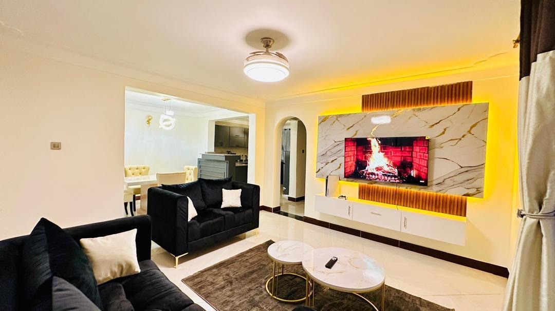 A modern living area features plush black sofas and a white marble fireplace, which is backlit with soft yellow light. An elegant round coffee table sits in the center, and a rug adds warmth to the space. A dining area is visible in the background.
