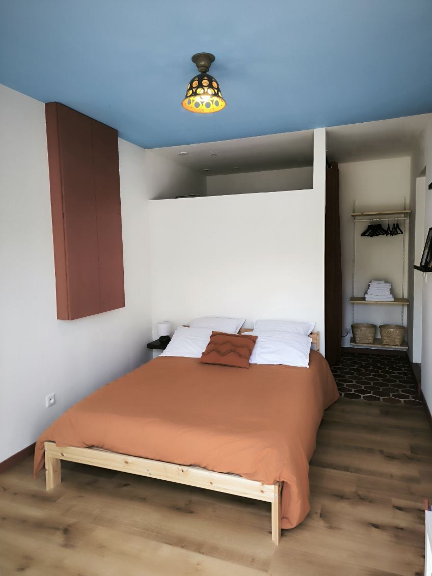 A comfortable sleeping area features a cozy bed with orange bedding and white pillows, positioned against a light wall. A bedside lamp and a small table are present on one side, while a wardrobe and storage shelves are visible in the background.