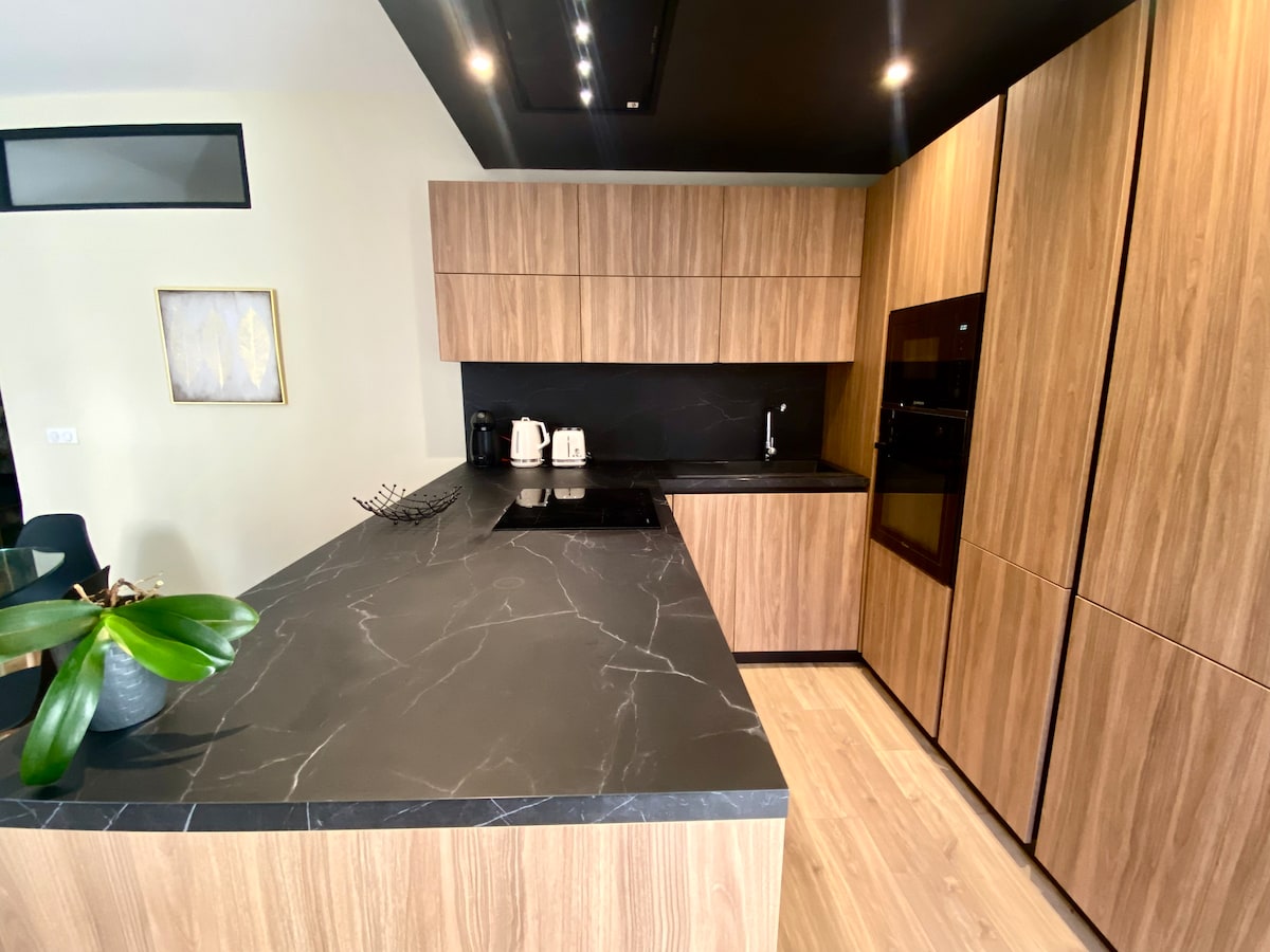 The modern kitchen features a large island with a dark countertop, providing ample preparation space. Wood cabinetry creates a warm atmosphere, complemented by sleek black appliances. A potted plant adds a touch of greenery, enhancing the kitchen's inviting design.