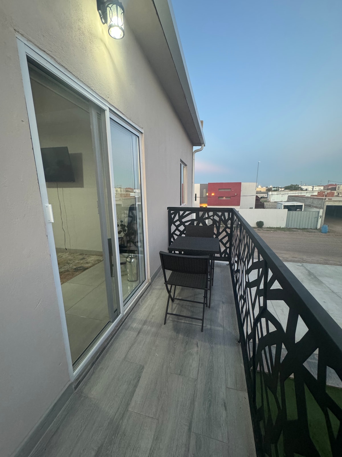 A balcony is featured with a black metal railing, offering a small table and two chairs for outdoor seating. The flooring is gray and appears even, extending the space. Large glass doors provide access to the interior while offering a view of the surroundings.