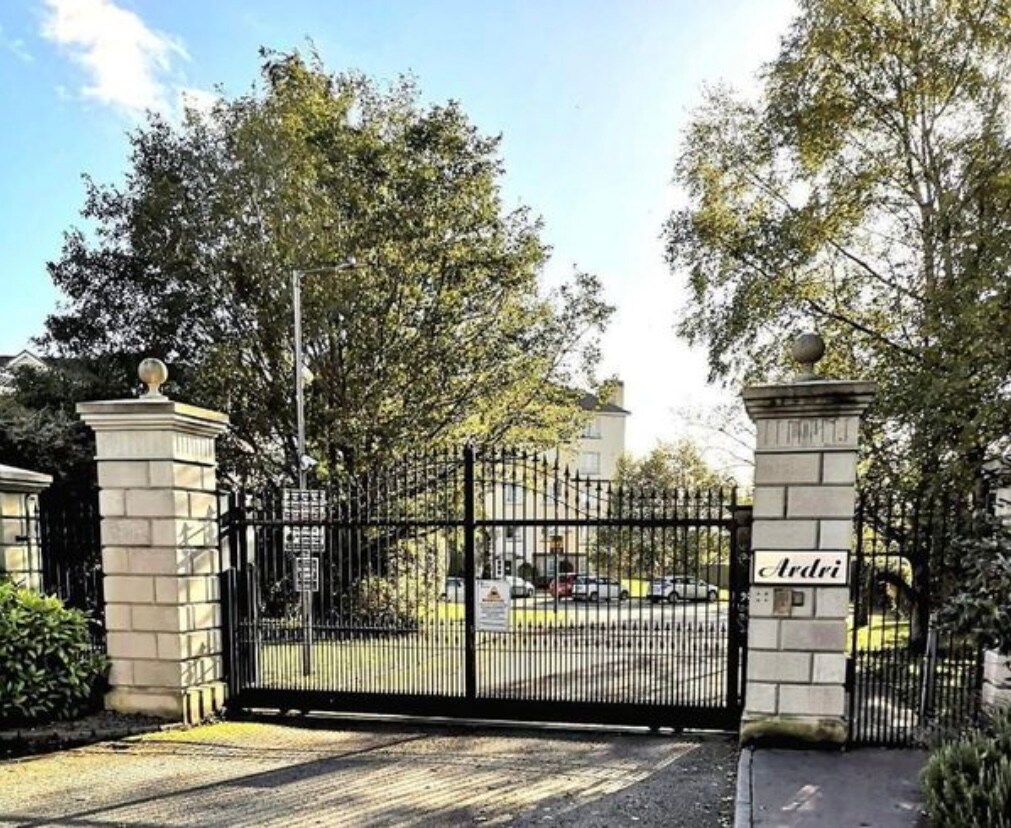A gated entrance is framed by tall stone pillars, each topped with decorative spheres. Lush green trees surround the entrance, providing a natural backdrop. A sign displaying the name 'Ardri' is prominently featured on one of the pillars.